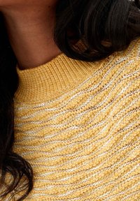 Gros plan sur une personne portant un pull en maille texturée jaune et blanc, montrant l'encolure et des cheveux ondulés foncés tombant sur l'épaule.