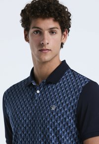 Navy poloshirt met een geometrisch driehoekpatroon in blauwtinten, met een effen navy kraag en een klein pinguïnlogo op de borst.