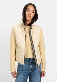 Beige quiltet puffer vest med høj krave, frontlynlås, to sidelommer og kontrasterende brun lynlåsdetalje, båret over en beige sweater.