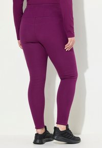 Person klädd i åtsittande lila leggings och svarta sportskor, stående mot en vit vägg med händerna längs sidorna.