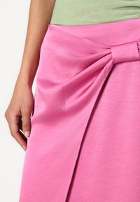 Pink skirt made of soft fabric, featuring a knotted waist detail and draped design. Smooth texture with a single hand holding the fabric.