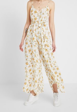 Femme portant une combinaison blanche sans manches à imprimé floral, jambes larges et baskets blanches sur fond uni.