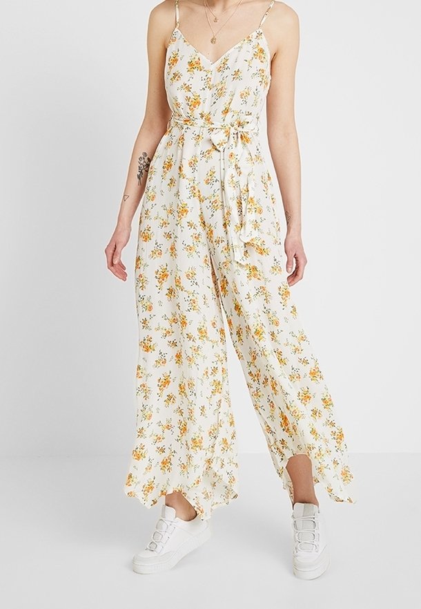Femme portant une combinaison blanche sans manches à imprimé floral, jambes larges et baskets blanches sur fond uni.