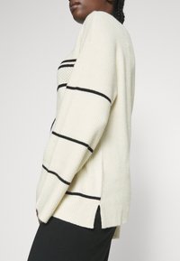 Ljus beige tröja med svarta horisontella ränder, ribbat mönster, sidoflikar och en avslappnad passform. Tillverkad av ett mjukt stickat material.