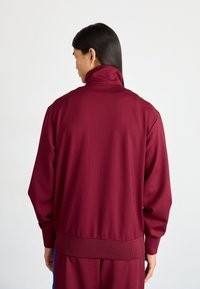 Giacca sportiva bordeaux con colletto alto, polsini e vita a costine. Texture liscia con vestibilità rilassata, caratterizzata da un motivo sottile.
