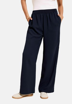 Osoba ubrana w szerokie granatowe spodnie z elastycznym pasem i białe tenisówki, z rękami w kieszeniach, stojąca na tle jednolitego tła.