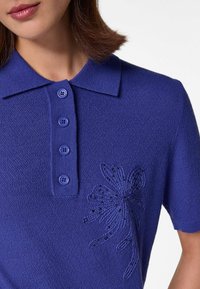 Lila Poloshirt mit strukturiertem Strick, einem Kragen und fünf passenden Knöpfen. Ein florales besticktes Design mit Verzierungen befindet sich auf der Brust.