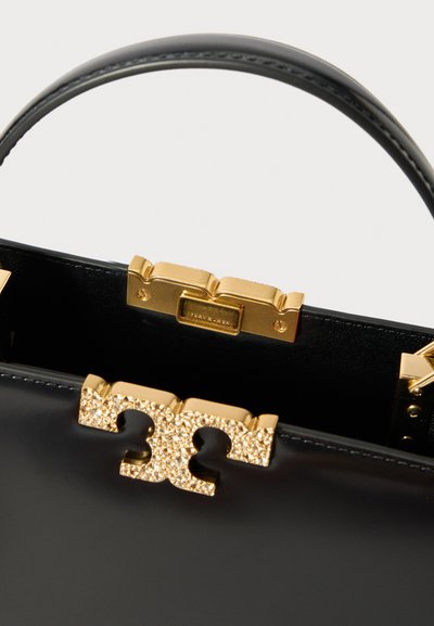 Melnas ādas rokassomiņa ar zeltainu spīdīgu aizdari un redzamu zelta metāla rāmi iekšpusē, ar izliekta ādas rokturi uz balta fona.