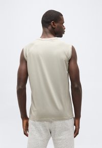 Άνδρας που στέκε ται με την πλάτη, φορώντας αμάνικη μπεζ μπλούζα και ανοιχτόχρωμο γκρι σορτς, μπροστά σε απλό φόντο.