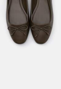 Un paio di ballerine in pelle marrone scuro con punta arrotondata e decorativi fiocchi sulla parte superiore, presentate su uno sfondo bianco.