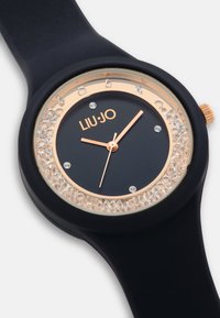 Orologio in silicone nero con cassa rotonda, dettagli in oro rosa, un quadrante abbellito con cristalli e un quadrante scuro con tre lancette e il logo.