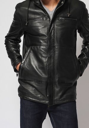 Osoba ubrana w czarną skórzaną kurtkę z kieszeniami na piersi zapinanymi na zamek oraz rękami wsuniętymi w boczne kieszenie, w połączeniu z ciemnoniebieskimi dżinsami.