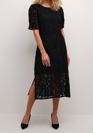 Kvinde iført en sort midi-kjole med blomstret blonder, puffærmer, sidelåg og sorte spidse hæle mod en ensfarvet baggrund.