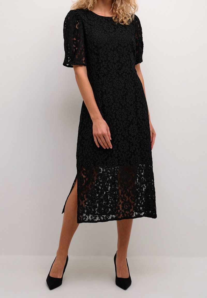 Femme portant une robe midi en dentelle noire à fleurs avec des manches bouffantes, une fente latérale, et des talons noirs à bout pointu, sur un fond uni.