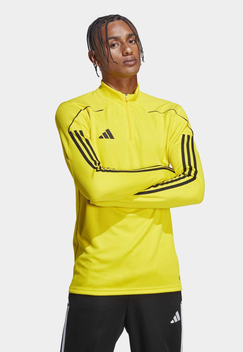Haut de sport long à manches jaunes avec col zippé, présentant des rayures noires sur les manches et un logo Adidas sur la poitrine. Texture de tissu lisse.