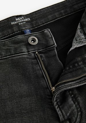 Zwarte slim-fit jeans met een metalen knop en ritssluiting. De stof heeft een gestructureerde afwerking met een subtiele glans. Binnenzijde van de tailleband met label zichtbaar.