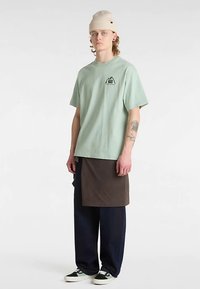 Mintgrøn t-shirt med korte ærmer og et bjerglogo, lagdelt med et brunt forklæde over marineblå vide bukser og sorte sneakers.