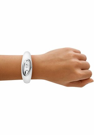 Poignet gauche portant un bracelet blanc et argenté avec un emblème métallique ressemblant à une languette de canette sur fond blanc.