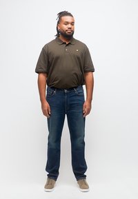 Άνδρας με ντραντλόκς που φοράει ελιά πόλο πουκάμισο, μπλε τζιν και μπεζ αθλητικά, στέκεται μπροστά σε λευκό φόντο.