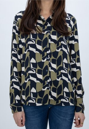 Vrouw draagt een blouse met lange mouwen in een abstract patroon van marineblauw, wit en olijfgroen, gecombineerd met een blauwe spijkerbroek.