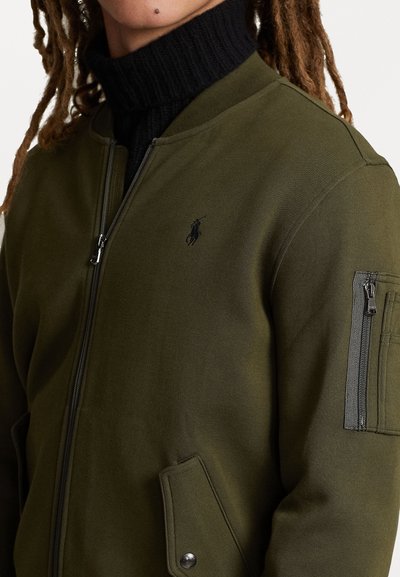 Casaco bomber verde-azeitona feito de tecido liso, com fecho de correr à frente, gola canelada e um pequeno logotipo no peito. Possui bolsos laterais e nas mangas.