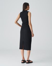 Schwarzes geripptes, ärmelloses Oberteil mit hohem Kragen, kombiniert mit einem Midirock mit Schlitz hinten. Das Model trägt flache schwarze Sandalen.