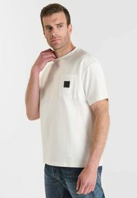 Uomo con capelli castani corti che indossa una maglietta bianca a maniche corte con una tasca sul petto e jeans scuri, in piedi contro un fondo neutro.