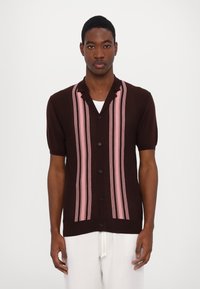 Hnědá krátká rukávová košile s růžovými vertikálními pruhy, s knoflíkovým zapínáním vpředu, kulatým límcem a texturovanou tkaninou. Nosená s bílými kalhotami.