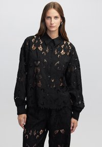 Camicia nera in pizzo con un motivo floreale, dotata di chiusura con bottoni e colletto. Maniche lunghe e tessuto trasparente conferiscono un aspetto intricato.