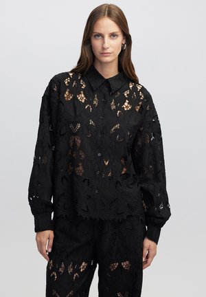 Chemise en dentelle noire ornée d'un motif floral, dotée d'une fermeture à boutons et d'un col. Les manches longues et la texture transparente confèrent un aspect raffiné.