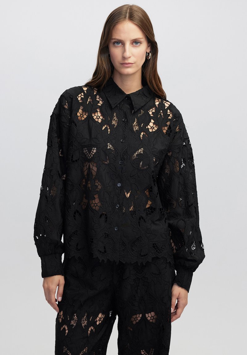 Zwarte kanten blouse met een bloemenpatroon, voorzien van een knoopsluiting en een kraag. Lange mouwen en een transparante textuur zorgen voor een ingewikkeld uiterlijk.
