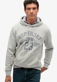 Lichtgrijze hoodie met buidelzak aan de voorkant, capuchon met trekkoord en bedrukte tekst. Voorzien van geribbelde boorden bij de mouwen en zoom, gecombineerd met blauwe spijkerbroek.
