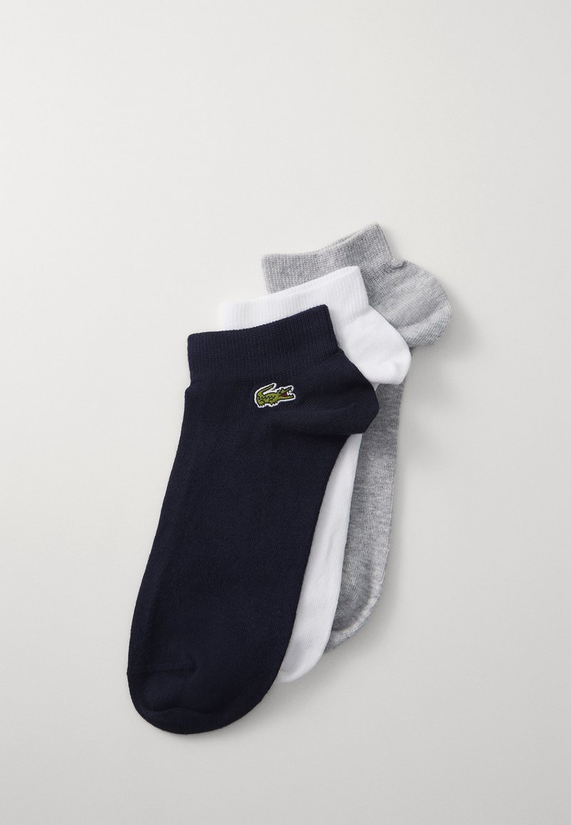 Drei Paare von Knöchelsocken: marineblau, weiß und hellgrau. Aus Baumwollmaterial mit geripptem Design und einem kleinen grünen Krokodil-Logo auf der marineblauen Socke.