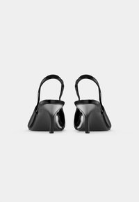 Saltos altos slingback pretos em verniz vistos de trás sobre um fundo branco liso.