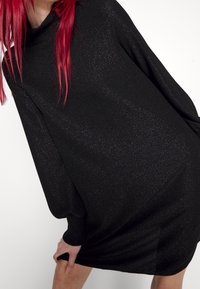 Robe-pull noire oversize en tissu texturé scintillant. Présente des manches longues et un col montant. Coupe fluide et décontractée.