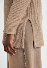 Pull à côtes beige avec fentes sur les côtés, présentant une texture douce. Main partiellement visible, mettant en avant les détails du tissu et le design global.