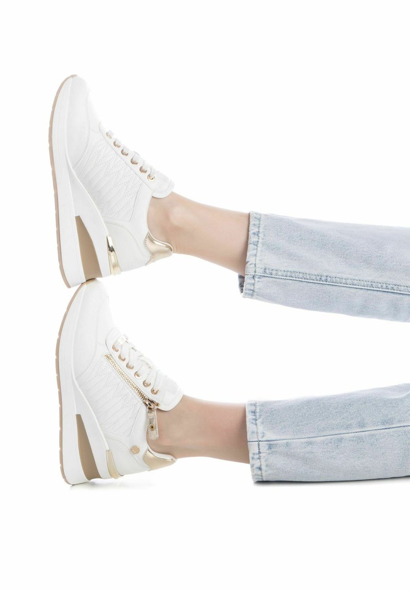 Zapatillas blancas con una parte superior texturizada, acentos dorados y una cremallera en el lateral. Tienen una suela con cuña y un diseño de cordones. Combinadas con jeans de mezclilla claros.