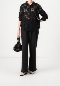 Svart spetsknäppt skjorta med geometriska mönster, kombinerad med vida svarta byxor. Håller en svart blommig dragsko-väska och bär beiga skor.