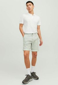 Witte polo shirt en lichtgroene shorts met opgerolde manchetten. Schoenen zijn grijs met zwarte accenten. Model staat met de handen in de zakken.
