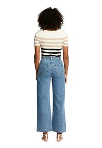 Top rayé côtelé en beige, noir et blanc ; assorti à un jean en denim bleu, taille haute et à jambes larges. La vue de derrière met en avant les détails subtils des poches.