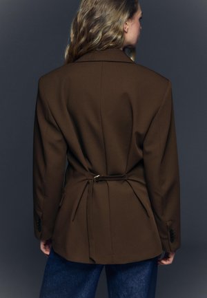 Frau mit welligem hellbraunem Haar, die einen dunkelbraunen, taillierten Blazer und dunkelblaue Jeans trägt, von hinten vor einem grauen Hintergrund.