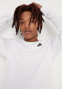 Valge pika varrukaga T-särk, mis on valmistatud puuvillast ja mille rinnal on must Nike logo, lõdva lõike ja ümmarguse kaelusega.