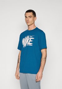 Blå bomulds t-shirt med et stort hvidt penselstrøg Nike-logo på fronten. Korte ærmer og rund halsudskæring, afslappet pasform.