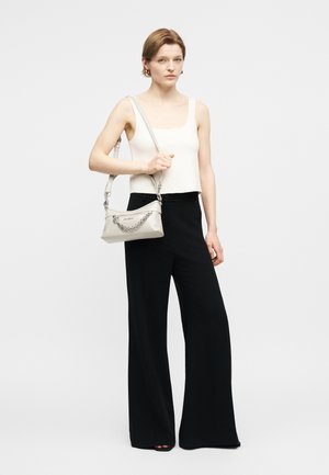 Jauna moteris su trumpais plaukais, apsirengusi balta be rankovių palaidine ir juodomis plačiomis kelnėmis, laikanti baltą petinę rankinę su grandinėlės detale.