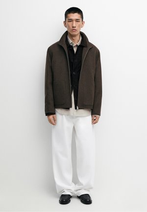 Mand iført lag-på-lag afslappet tøj, inklusive en brun jakke, sort skjorte, beige skjorte, hvide bukser og sorte sko, står mod en hvid baggrund.