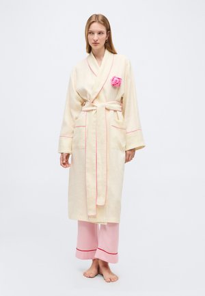 Žena v krémovom župane s ružovým lemovaním a opaskom, s brošňou v tvare ružovej ruže na hrudi a svetloružových pyžamových nohaviciach s červenými manžetami, stojaca naboso.