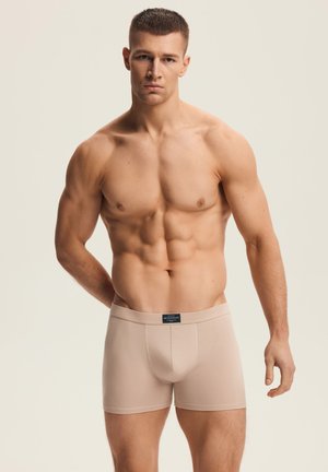Muskulöser Mann mit kurzem Haar, der beigen Boxer-Shorts trägt und vor einem schlichten hellen Hintergrund steht, mit dem Gesicht nach vorn und einem Arm hinter seinem Rücken.