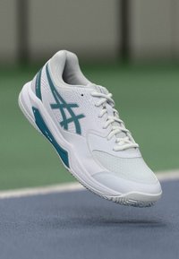 Weißer Sportschuh mit Türkisakzenten, strukturiertem Mesh-Obermaterial, gepolstertem Kragen, Schnürsenkeln und einer Gummisohle mit einem gemusterten Profil.