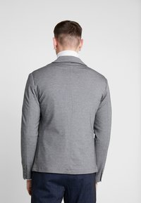 Blazer grigio con texture, colletto a punta, design aderente e dettagli con bottoni. Vista posteriore, abbinato a pantaloni navy a fantasia.
