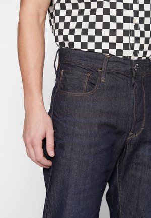 Personne portant un jean en denim bleu foncé et une chemise à carreaux noir et blanc, la main détendue le long du corps.